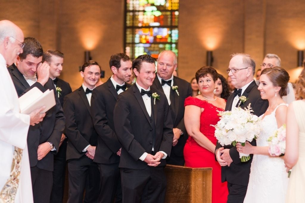 Bride in a lace wedding gown holding a white bouquet during a traditional church ceremony, standing with the groom, groomsmen in black tuxedos, and family members as the officiant leads the wedding service.