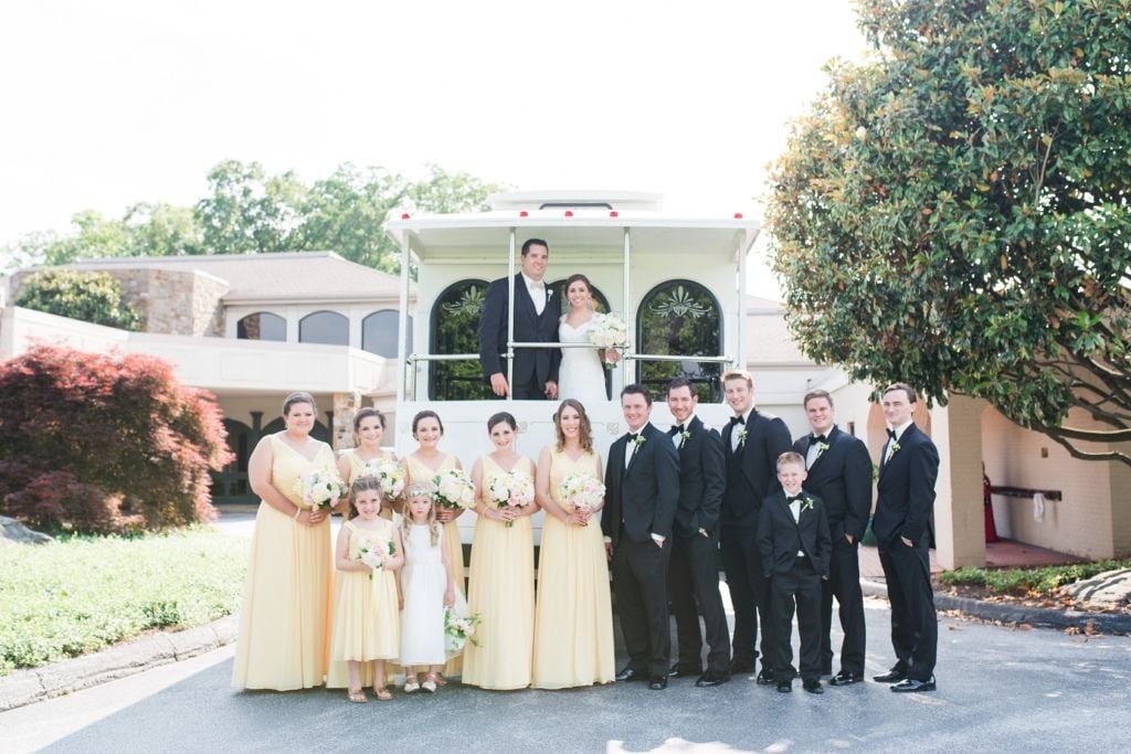 Bride and groom standing on a white trolley with their wedding party outdoors, featuring bridesmaids in soft yellow dresses holding bouquets and groomsmen in classic black tuxedos, captured during a formal wedding portrait by K&B Bridals.