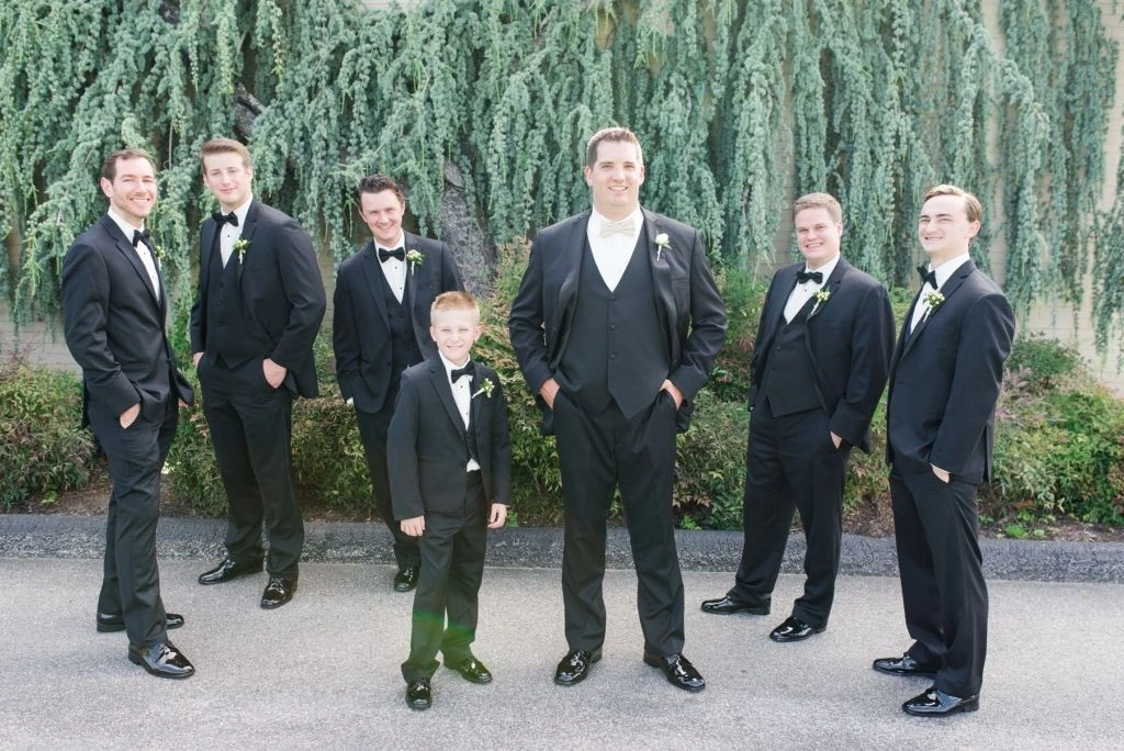 Group portrait of the groom and groomsmen dressed in black tuxedos, standing outdoors in front of lush greenery with relaxed, smiling poses.
