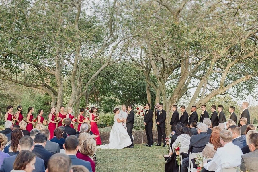 Bride and groom standing at the altar during an outdoor wedding ceremony, surrounded by bridesmaids in red dresses and groomsmen in black suits under large trees with seated guests watching.