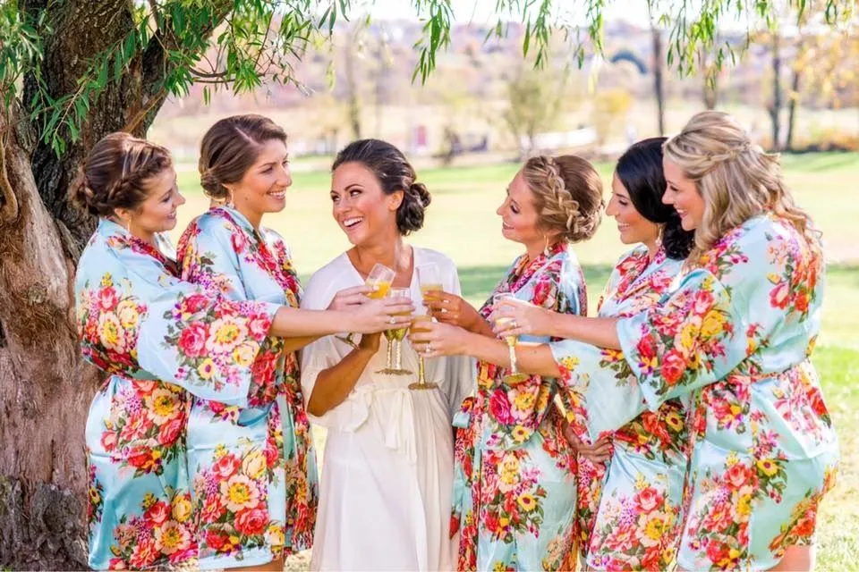Bride and her bridesmaids wearing colorful floral robes, standing outdoors under a tree and raising champagne glasses together during a joyful pre-wedding celebration.