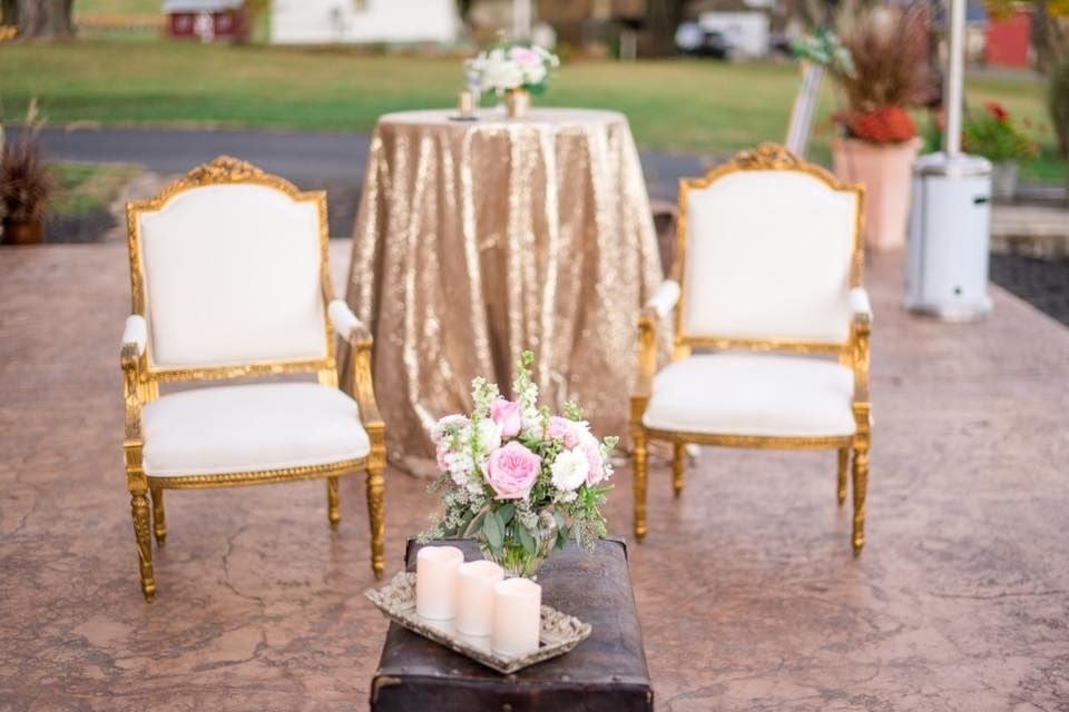 Outdoor wedding ceremony setup featuring two ornate gold chairs, a floral arrangement with pink roses, candles, and a gold-draped table, styled for a romantic wedding celebration by K&B Bridals.
