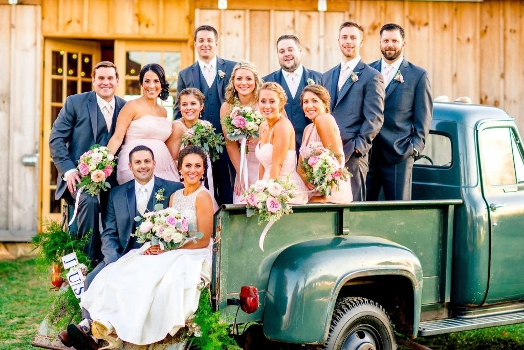 Bride, groom, bridesmaids in blush dresses, and groomsmen in gray suits posing together around a vintage green pickup truck for a joyful outdoor wedding party photo.