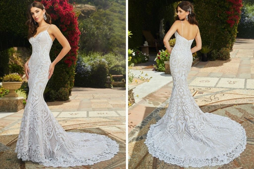 Sneak Peek: Casablanca Bridal Tester Event Front and back views of a lace fit-and-flare wedding gown featuring bold geometric and floral patterns, spaghetti straps, and an ornate scalloped train, photographed outdoors in a garden setting.