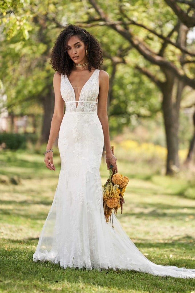 Bride wearing a lace sheath wedding dress with a deep V-neckline, illusion plunge detail, and soft train, styled outdoors in a garden setting with natural greenery and a textured floral bouquet.