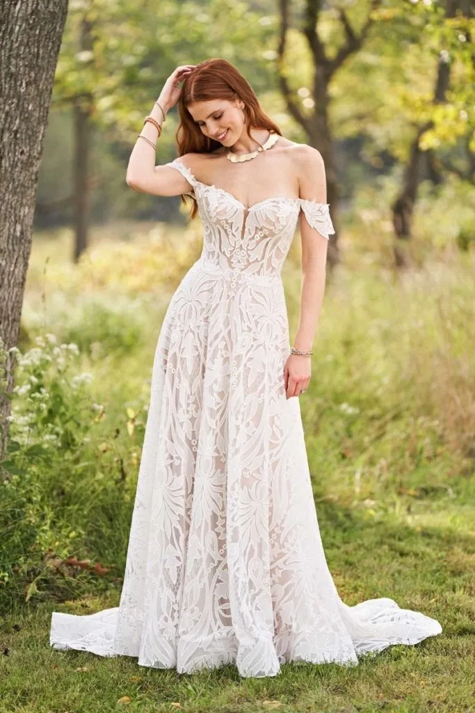 Bride wearing an off-the-shoulder lace A-line wedding dress with a sweetheart neckline and intricate floral detailing, standing in a sunlit woodland field.