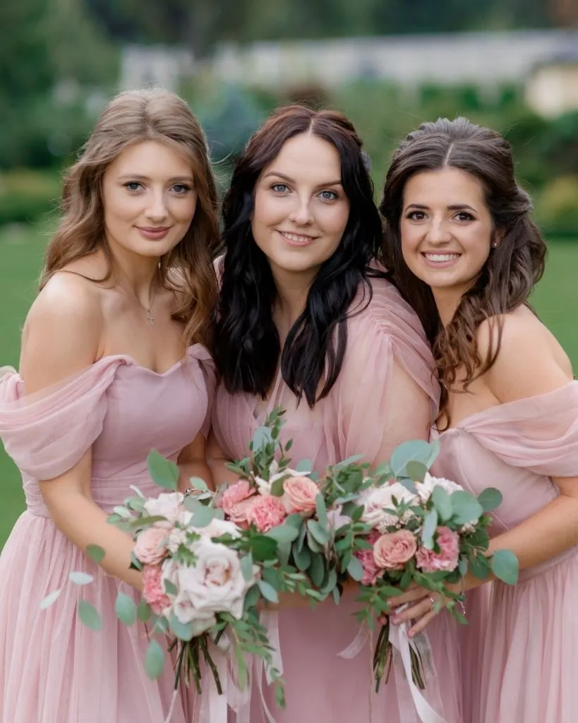 Traveler’s Club: 3 Unforgettable Getaways You Can Unlock with Your Wedding Dress Purchase Three bridesmaids wearing blush pink off-the-shoulder dresses, holding matching bouquets of pink and white flowers with eucalyptus greenery, smiling together outdoors.