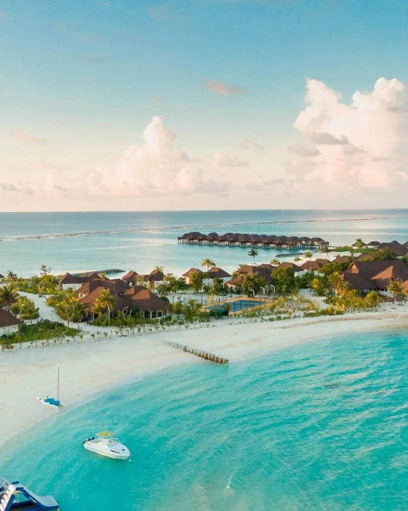 Traveler’s Club: 3 Unforgettable Getaways You Can Unlock with Your Wedding Dress Purchase Aerial view of a tropical beachfront resort with turquoise water, sandy shores, boats, and rows of overwater villas extending into the ocean under a bright sky.