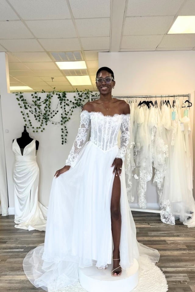 Page Updates & Announcements Bride modeling a romantic off-the-shoulder wedding gown with lace sleeves, corset bodice, and a flowing tulle skirt with a high slit.