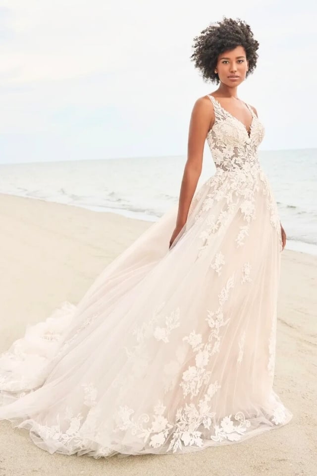 Bride on beach wearing sleeveless A-line wedding gown with lace appliqués, deep V-neckline, and long flowing train.