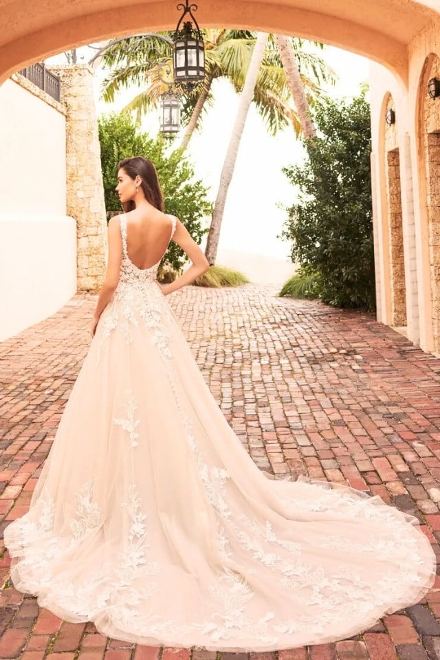 Elegant Floral A-Line Wedding Dress Tillie MT3210 Bride in sleeveless A-line wedding dress with deep V-neckline, floral lace detailing, and long glitter tulle train, standing on cobblestone path.