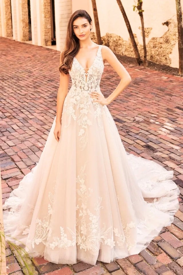 Bride in sleeveless A-line wedding dress with deep V-neckline, floral lace detailing, and long glitter tulle train, standing on cobblestone path.