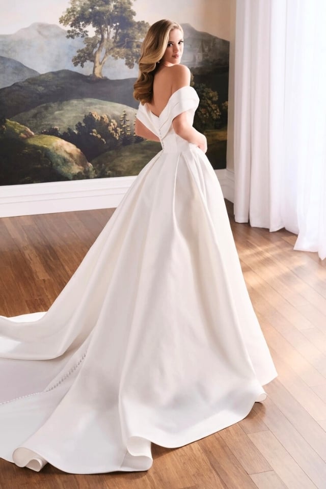 Off-Shoulder Mikado Ball Gown Wedding Dress Wren MT8112 Bride in off-shoulder Mikado ball gown with ruched bodice and full pleated skirt in Diamond White, standing against a vintage landscape mural backdrop.