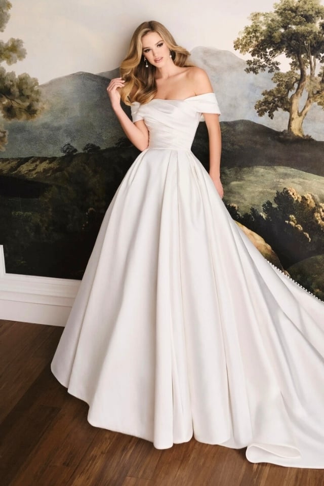 Bride in off-shoulder Mikado ball gown with ruched bodice and full pleated skirt in Diamond White, standing against a vintage landscape mural backdrop.