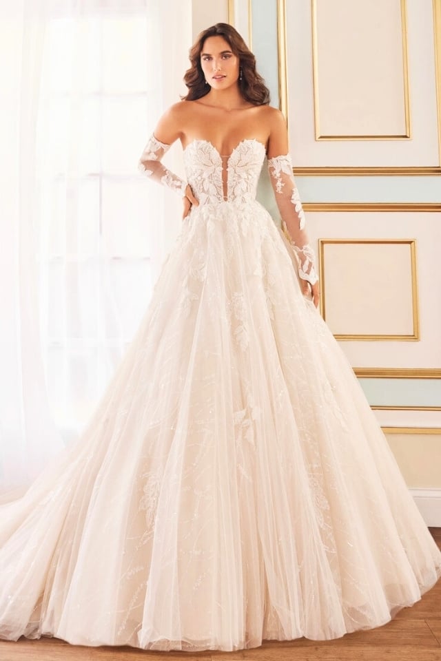 Bride in sparkling tulle ball gown with sweetheart neckline and lace embroidery, wearing off-the-shoulder sleeves and posing in a soft-lit, elegant room.