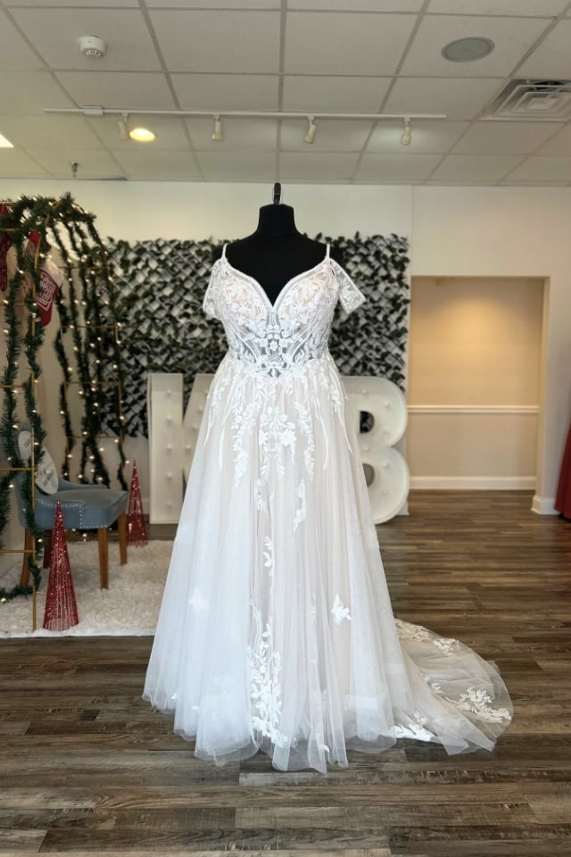 A-line wedding gown with off-the-shoulder sleeves, V-neckline, and floral lace appliqués on a sparkly tulle skirt displayed on mannequin in bridal boutique.