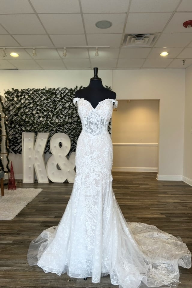 Off-the-shoulder lace wedding gown with sweetheart neckline, floral appliqués, and soft tulle train displayed on a mannequin in bridal boutique.