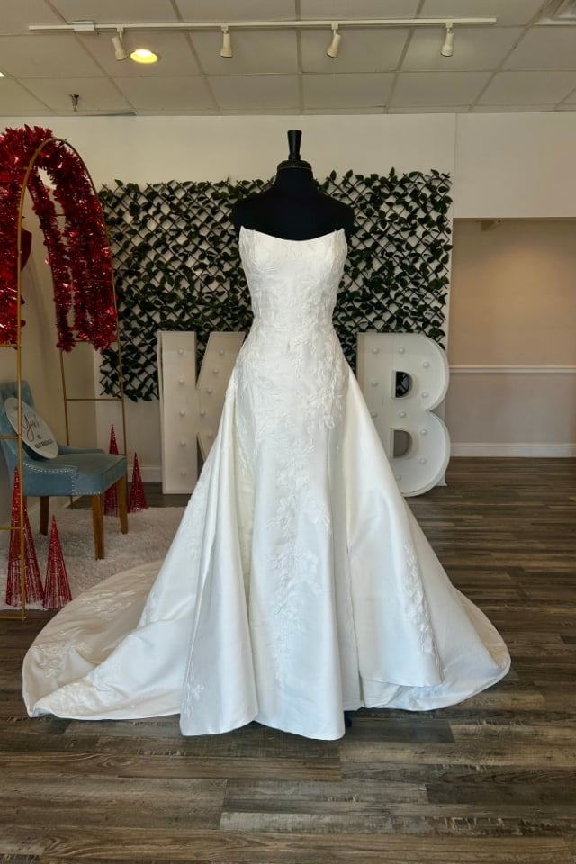 Page Updates & Announcements Strapless satin A-line wedding dress with floral lace appliqués displayed on mannequin in bridal boutique.
