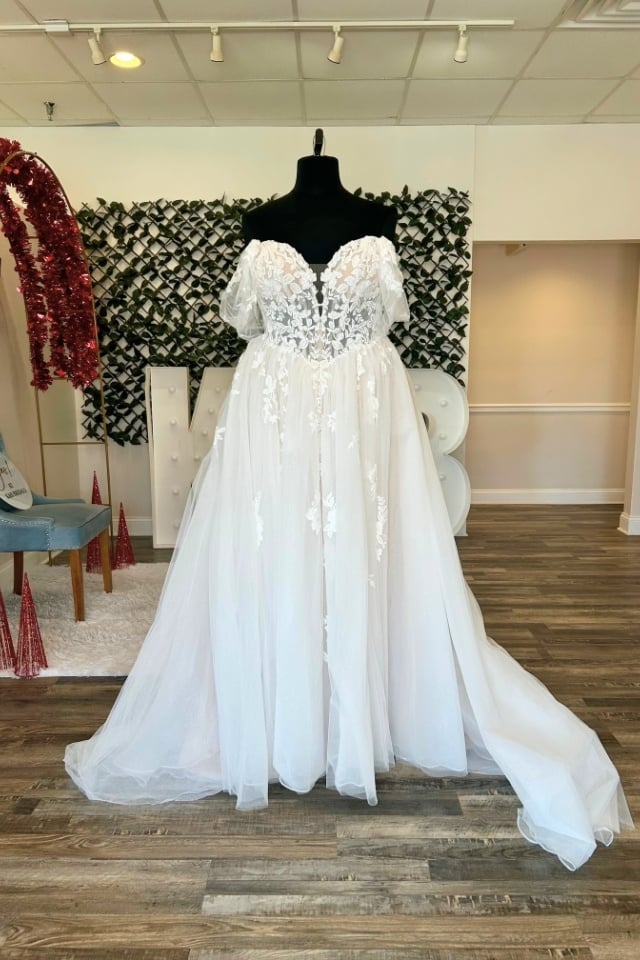 Page Updates & Announcements Off-the-shoulder lace A-line wedding dress with floral appliqués and glitter tulle skirt displayed on mannequin in bridal boutique.
