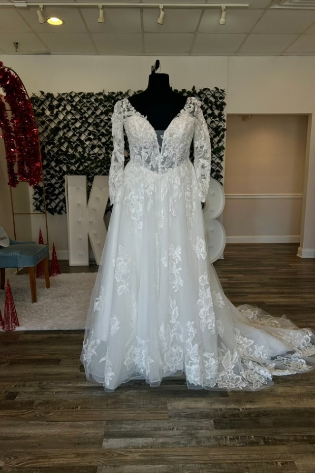 Page Updates & Announcements Long sleeve lace A-line wedding dress with V-neck illusion bodice and floral appliqués displayed on mannequin in bridal boutique.