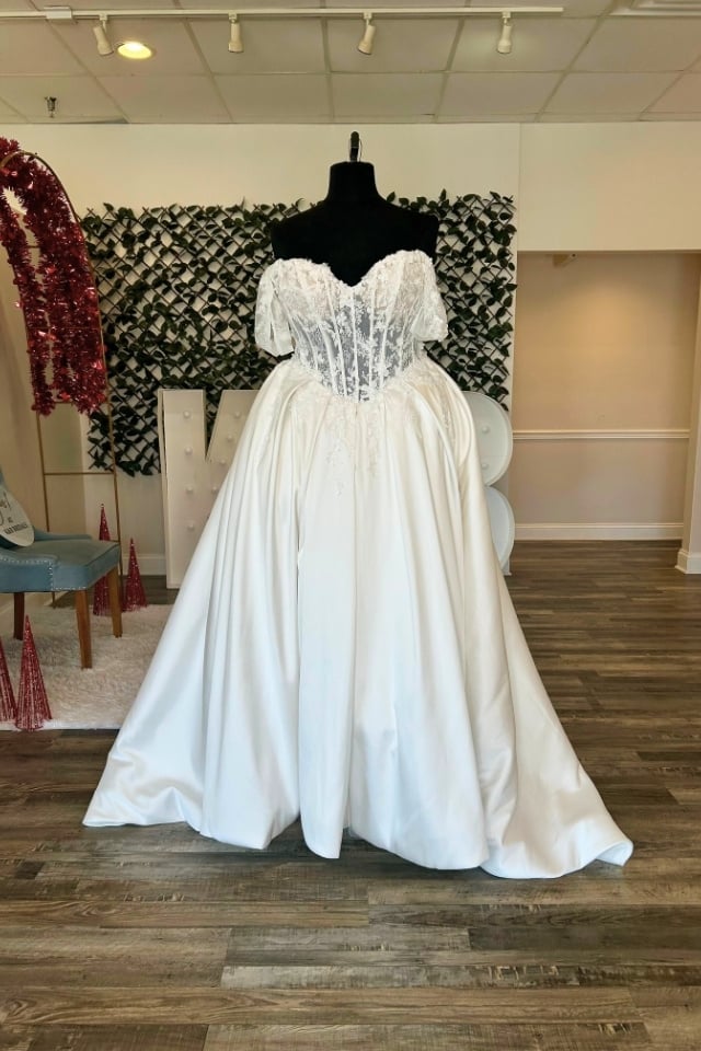 Off-the-shoulder lace and satin ball gown wedding dress with corset bodice and full skirt displayed on mannequin in bridal boutique.