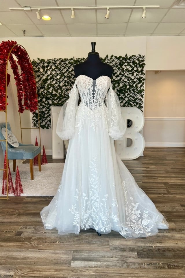 Off-the-shoulder lace A-line wedding dress with sheer sleeves and floral appliqués displayed on mannequin in bridal boutique.