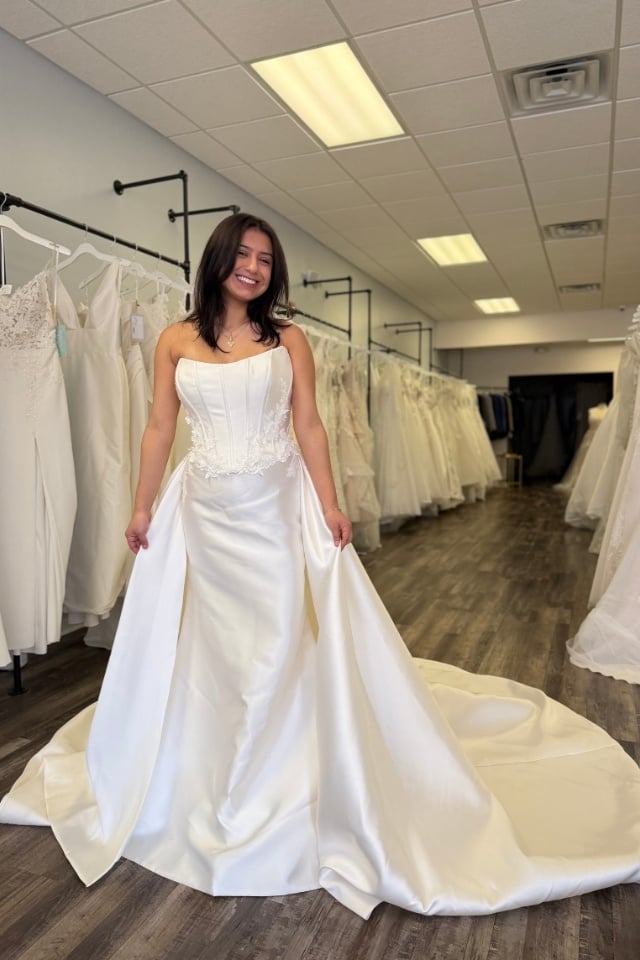 Convertible satin wedding dress with corset bodice and detachable overskirt worn by bride in bridal boutique.