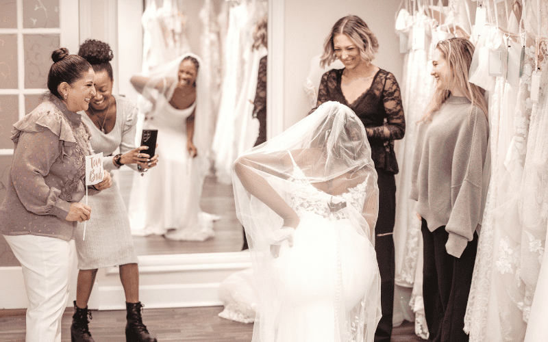 How to Find the Perfect Wedding Dress Beyond Your Pinterest Board woman with stylist and friends in front of the mirror laughing wearing a wedding dress
