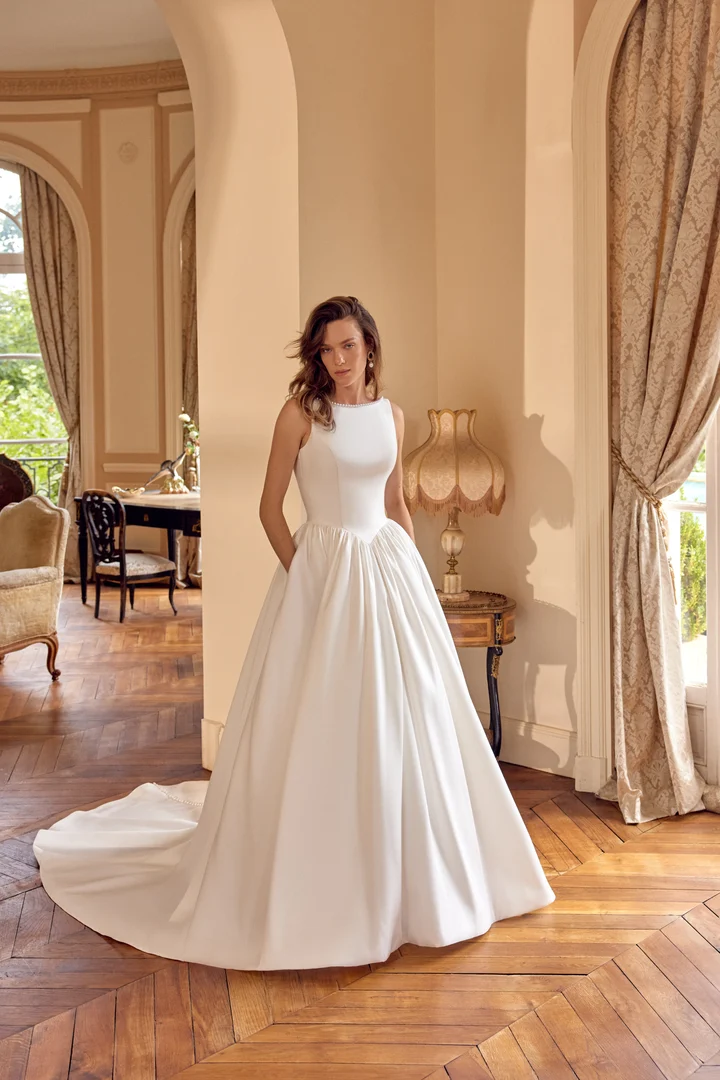 McKenna Mikado ball gown wedding dress featuring a pearl-trimmed Sabrina neckline, sleeveless fitted bodice, basque waistline, full pleated skirt with pockets, V-shaped back, covered buttons, and chapel-length train.