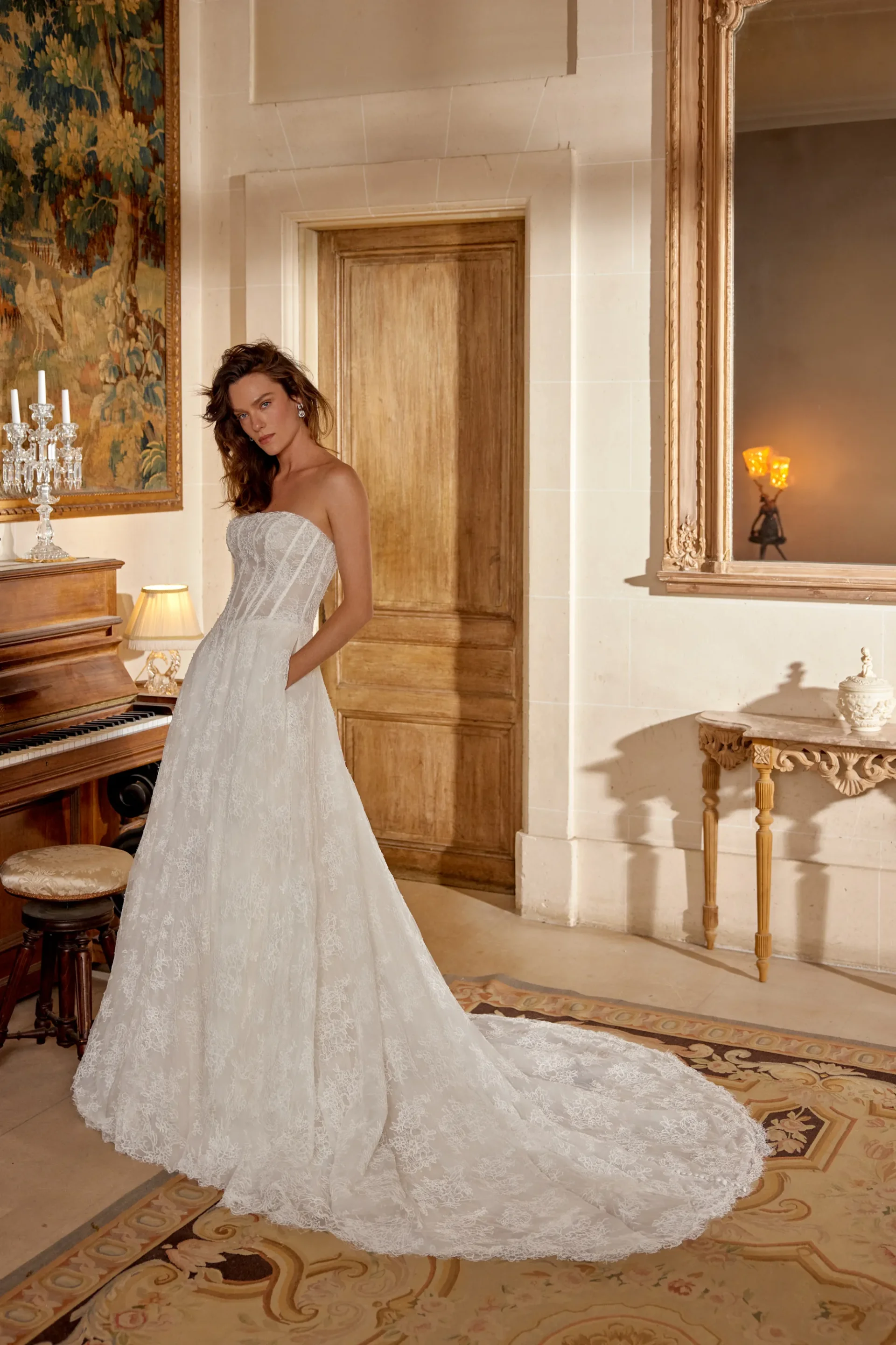 Manuela by Justin Alexander ivory lace A-line wedding dress featuring a strapless straight neckline, structured corset bodice, basque waistline, flowing lace skirt, side pockets, and chapel-length train.