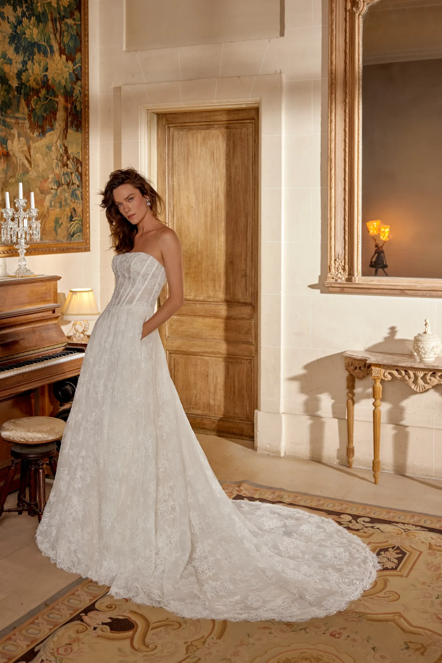 Strapless Lace A-Line Wedding Dress Manuela Manuela by Justin Alexander ivory lace A-line wedding dress featuring a strapless straight neckline, structured corset bodice, basque waistline, flowing lace skirt, side pockets, and chapel-length train.